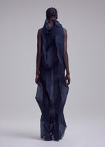 With short, braided hair and facing away, the person wears ISSEY MIYAKE’s SHADE AND SHADED_NY: a sheer dark blue long dress with geometric patterns and layered, sculptural fabric, set against a plain light gray background.