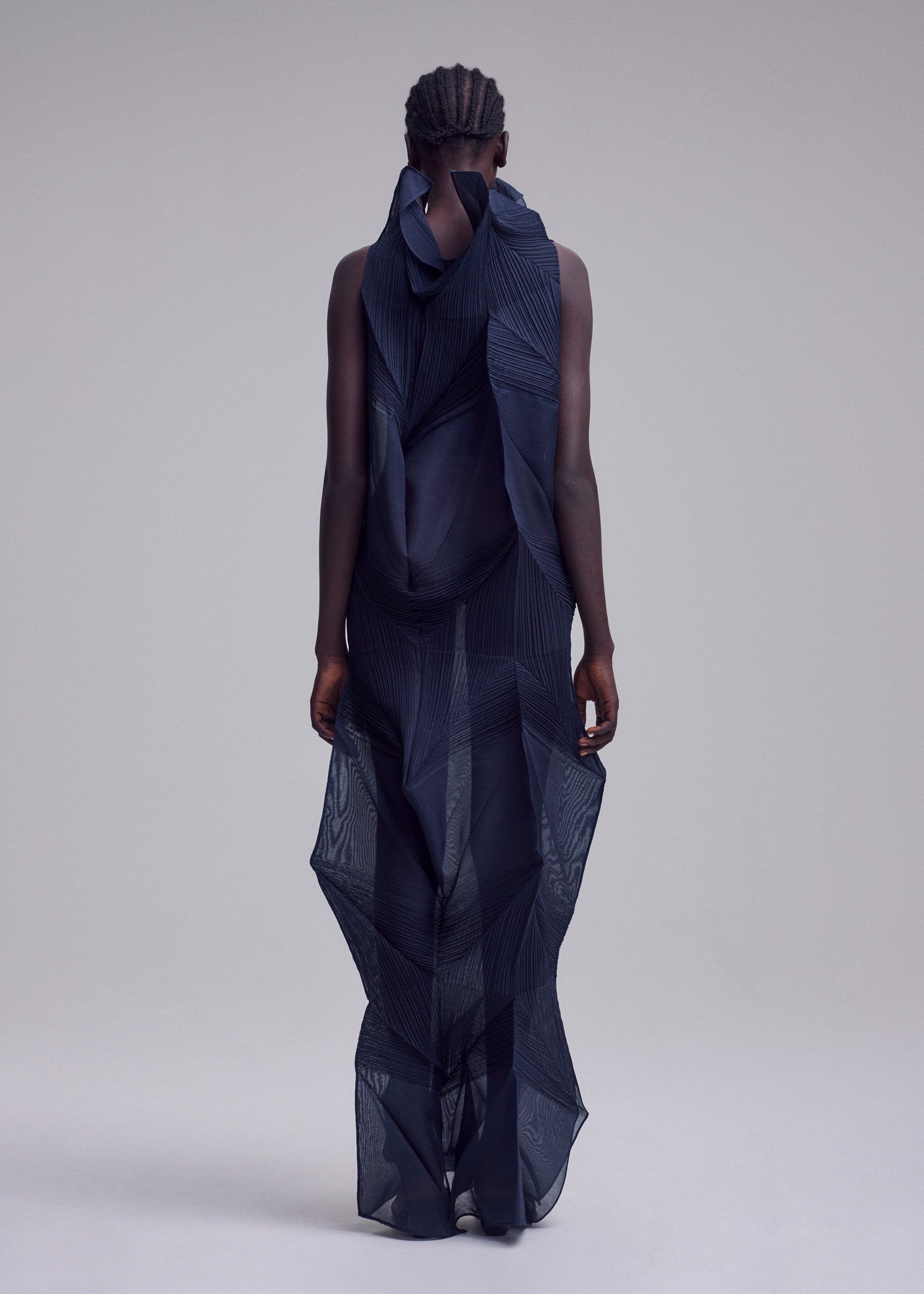 Zoomed view With short, braided hair and facing away, the person wears ISSEY MIYAKE’s SHADE AND SHADED_NY: a sheer dark blue long dress with geometric patterns and layered, sculptural fabric, set against a plain light gray background.