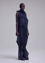 A person stands against a plain light background, wearing the SHADE AND SHADED_NY dress by ISSEY MIYAKE—a dark, machine-pleated, sleeveless long dress with sheer overlapping panels and a high structured collar.