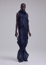 A person stands against a plain light gray background, wearing the ISSEY MIYAKE SHADE AND SHADED_NY dress—navy, sheer, machine-pleated with geometric patterns and wave-like layers—paired with flat sandals.