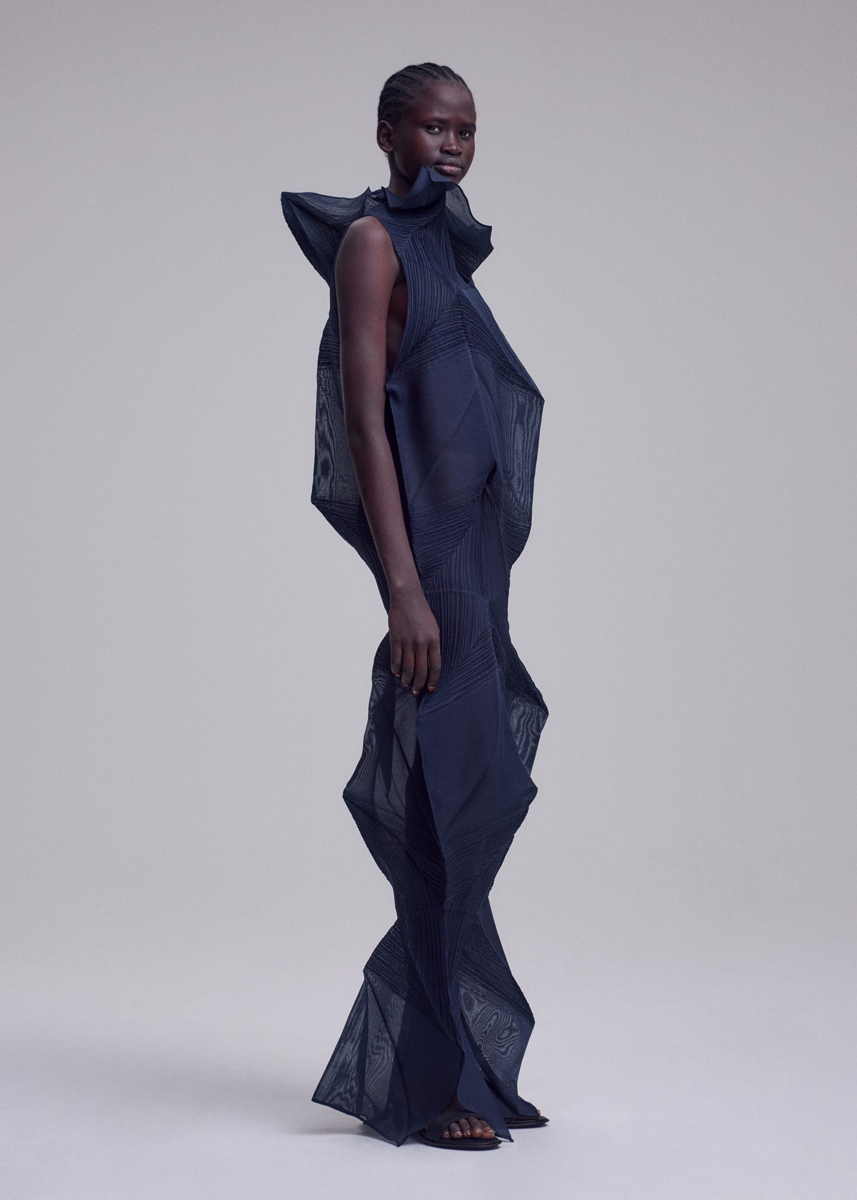 A model stands in profile wearing ISSEY MIYAKE’s SHADE AND SHADED_NY, a sculptural dark navy long dress with machine-pleated geometric shapes and textured patterns, set against a plain light gray background. view 10 of 10