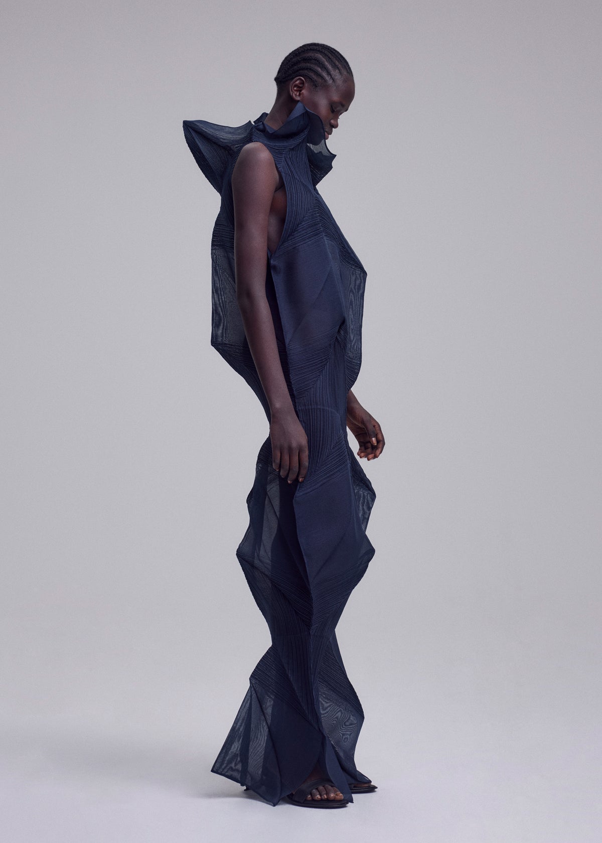 A person with short, braided hair wears the SHADE AND SHADED_NY machine-pleated long dress by ISSEY MIYAKE, featuring a sculptural geometric silhouette with pronounced shoulders, neck, and legs, set against a plain background. view 7 of 10