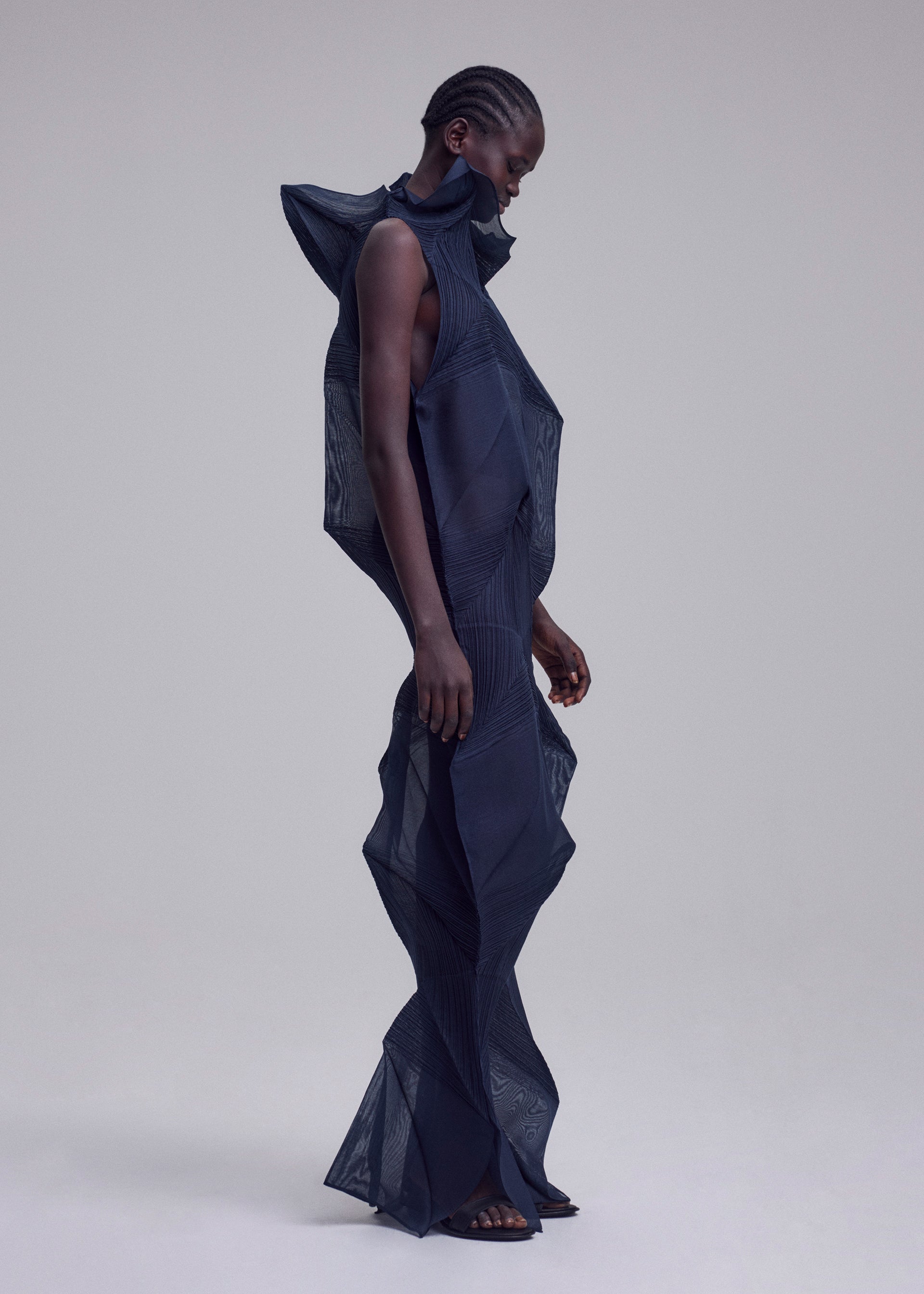 Zoomed view A person with short, braided hair wears the SHADE AND SHADED_NY machine-pleated long dress by ISSEY MIYAKE, featuring a sculptural geometric silhouette with pronounced shoulders, neck, and legs, set against a plain background.