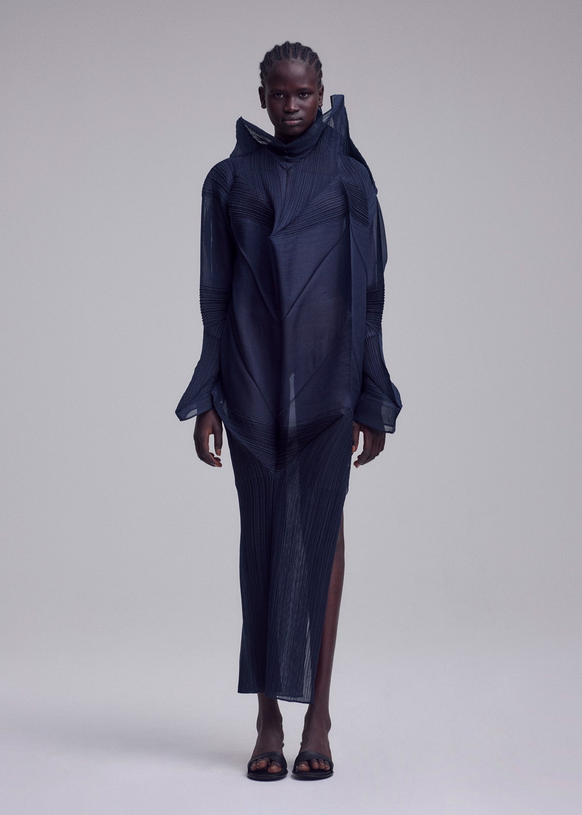 Someone poses against a plain background in the ISSEY MIYAKE SHADE AND SHADED_NY dress—a sheer, dark, sculptural design with structured lines, long sleeves, high neckline, skirt slit—and open-toe black sandals. view 3 of 10