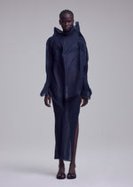 Someone poses against a plain background in the ISSEY MIYAKE SHADE AND SHADED_NY dress—a sheer, dark, sculptural design with structured lines, long sleeves, high neckline, skirt slit—and open-toe black sandals.