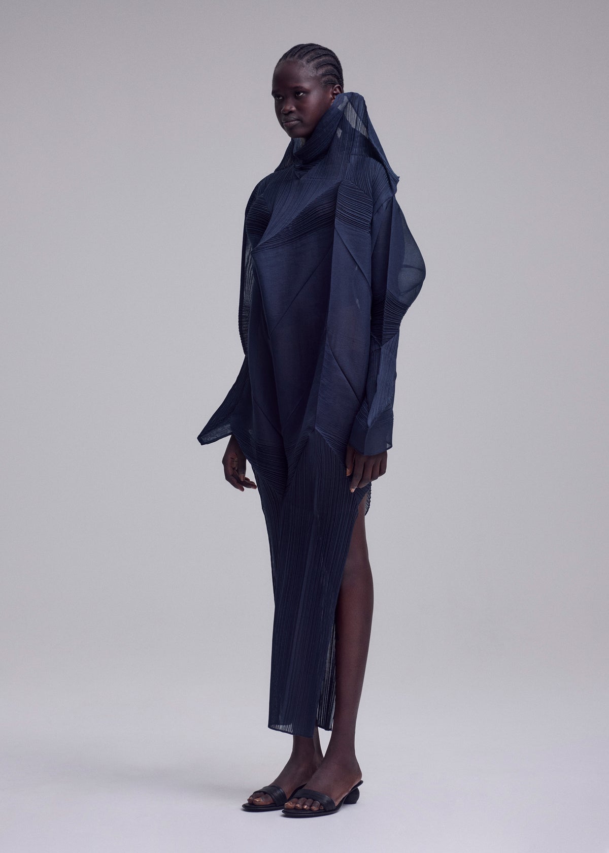 A person with dark braided hair wears the ISSEY MIYAKE SHADE AND SHADED_NY dress—sculptural, long-sleeved, and dark navy with geometric details and a high collar—paired with black sandals against a plain light grey background. view 4 of 10