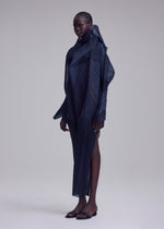 A person with dark braided hair wears the ISSEY MIYAKE SHADE AND SHADED_NY dress—sculptural, long-sleeved, and dark navy with geometric details and a high collar—paired with black sandals against a plain light grey background.