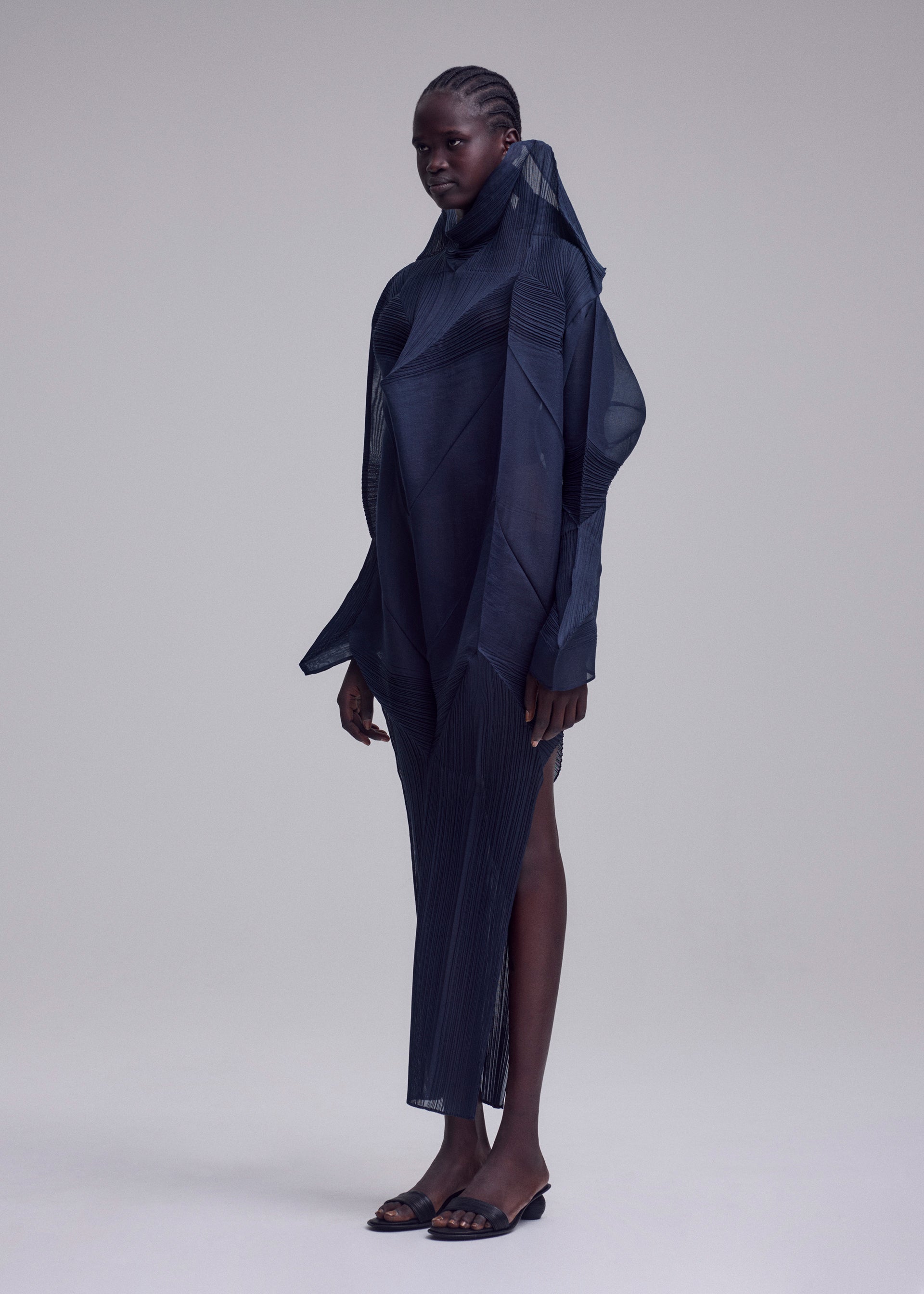 Zoomed view A person with dark braided hair wears the ISSEY MIYAKE SHADE AND SHADED_NY dress—sculptural, long-sleeved, and dark navy with geometric details and a high collar—paired with black sandals against a plain light grey background.