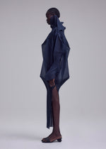 A person with dark skin and braided hair stands in profile, wearing ISSEY MIYAKE's SHADE AND SHADED_NY dress—a structured navy piece with sheer panels and pointed shoulders—paired with open-toed black sandals against a neutral background.