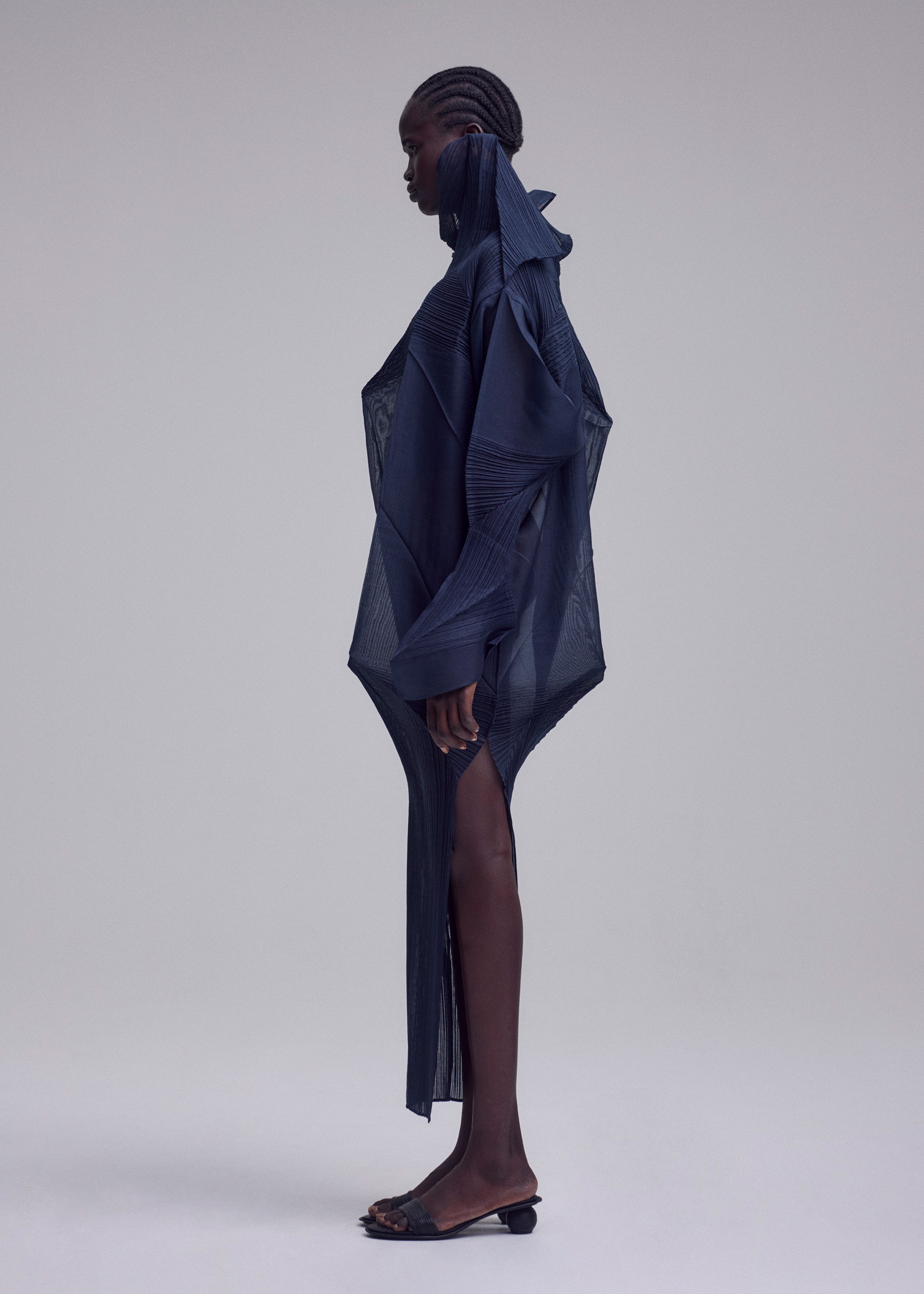 Zoomed view A person with dark skin and braided hair stands in profile, wearing ISSEY MIYAKE's SHADE AND SHADED_NY dress—a structured navy piece with sheer panels and pointed shoulders—paired with open-toed black sandals against a neutral background.