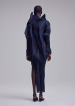 A person with short, braided hair stands facing away, wearing the SHADE AND SHADED_NY dress by ISSEY MIYAKE— a dark, sculptural piece with geometric shapes and sheer panels—paired with black low-heeled shoes against a plain background.