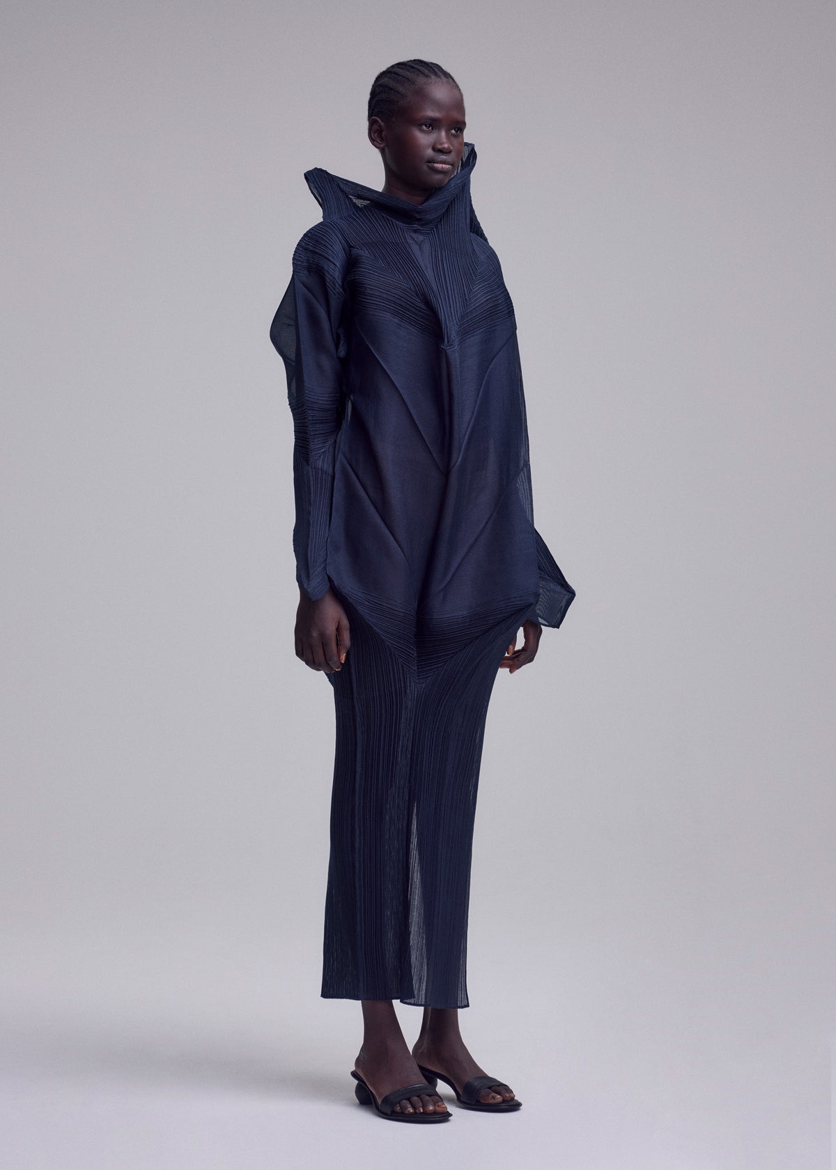 A person stands against a plain light background, wearing the SHADE AND SHADED_NY long-sleeved sculptural dress with geometric lines by ISSEY MIYAKE and open-toed sandals. Their hair is styled in tight braids pulled back. view 8 of 10