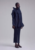 A person stands against a plain light background, wearing the SHADE AND SHADED_NY long-sleeved sculptural dress with geometric lines by ISSEY MIYAKE and open-toed sandals. Their hair is styled in tight braids pulled back.