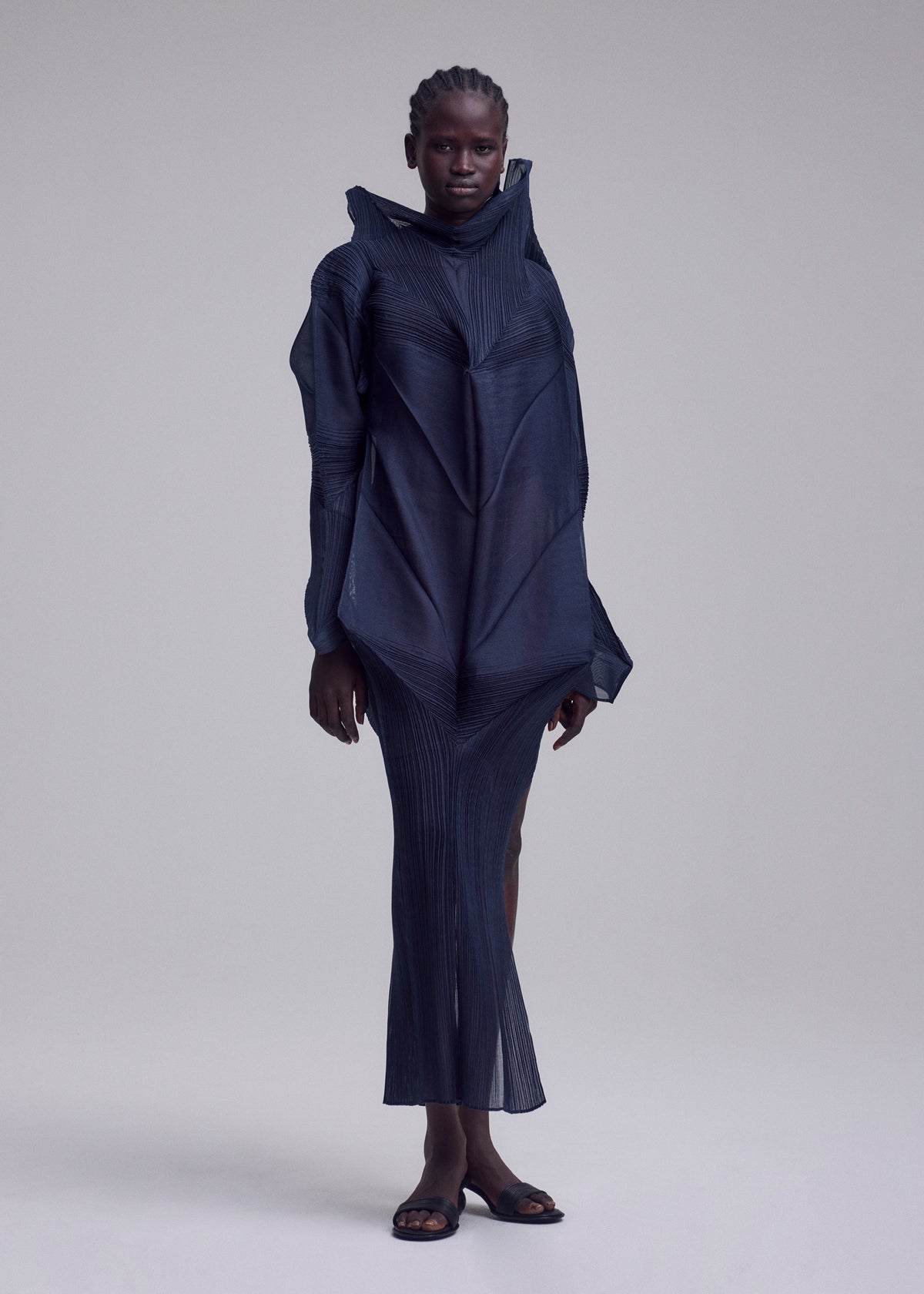 Against a plain background, a person with short, braided hair wears the SHADE AND SHADED_NY flowing pleated long-sleeved geometric dress by ISSEY MIYAKE, paired with sandals. view 9 of 10
