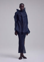 Against a plain background, a person with short, braided hair wears the SHADE AND SHADED_NY flowing pleated long-sleeved geometric dress by ISSEY MIYAKE, paired with sandals.