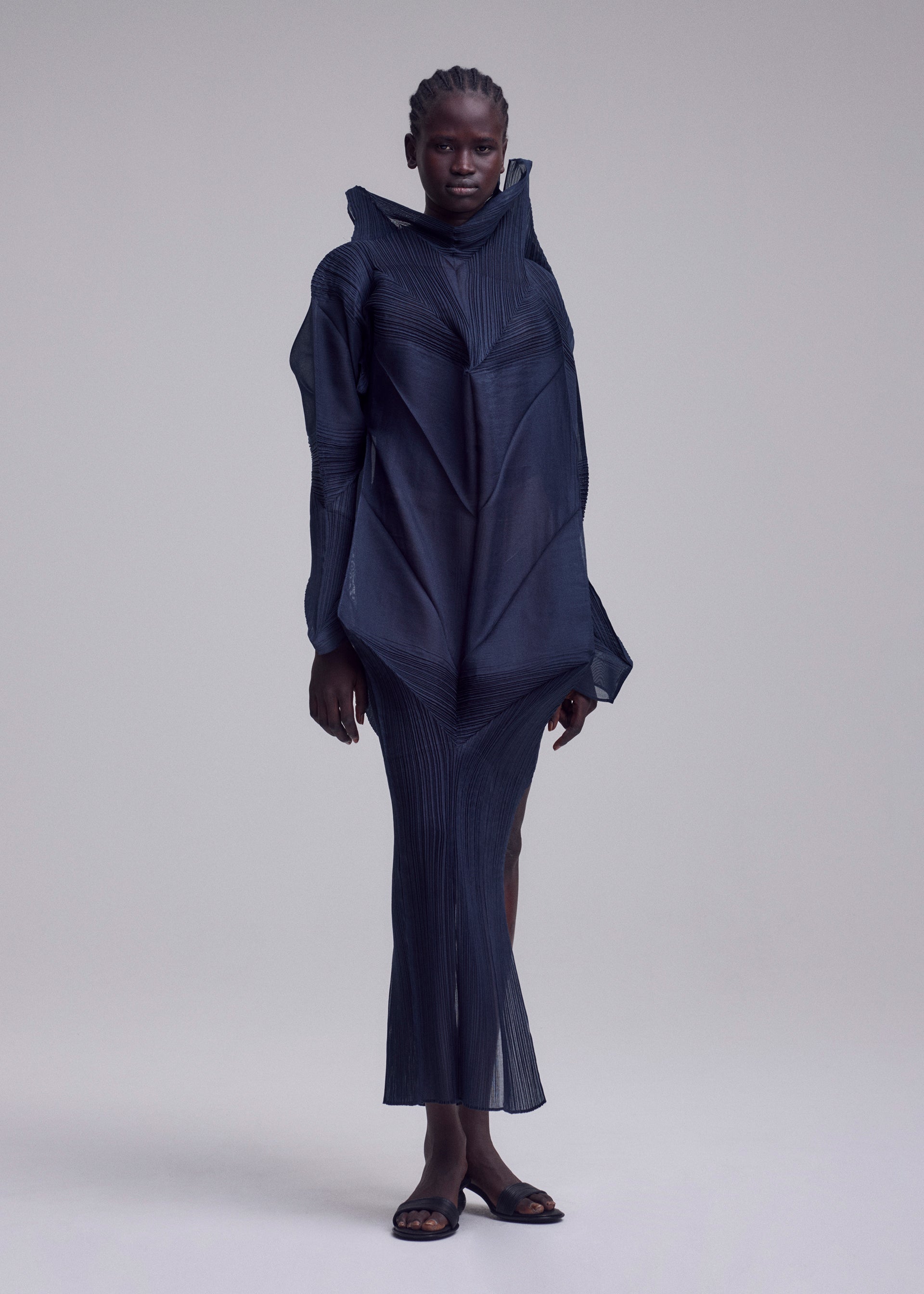 Zoomed view Against a plain background, a person with short, braided hair wears the SHADE AND SHADED_NY flowing pleated long-sleeved geometric dress by ISSEY MIYAKE, paired with sandals.
