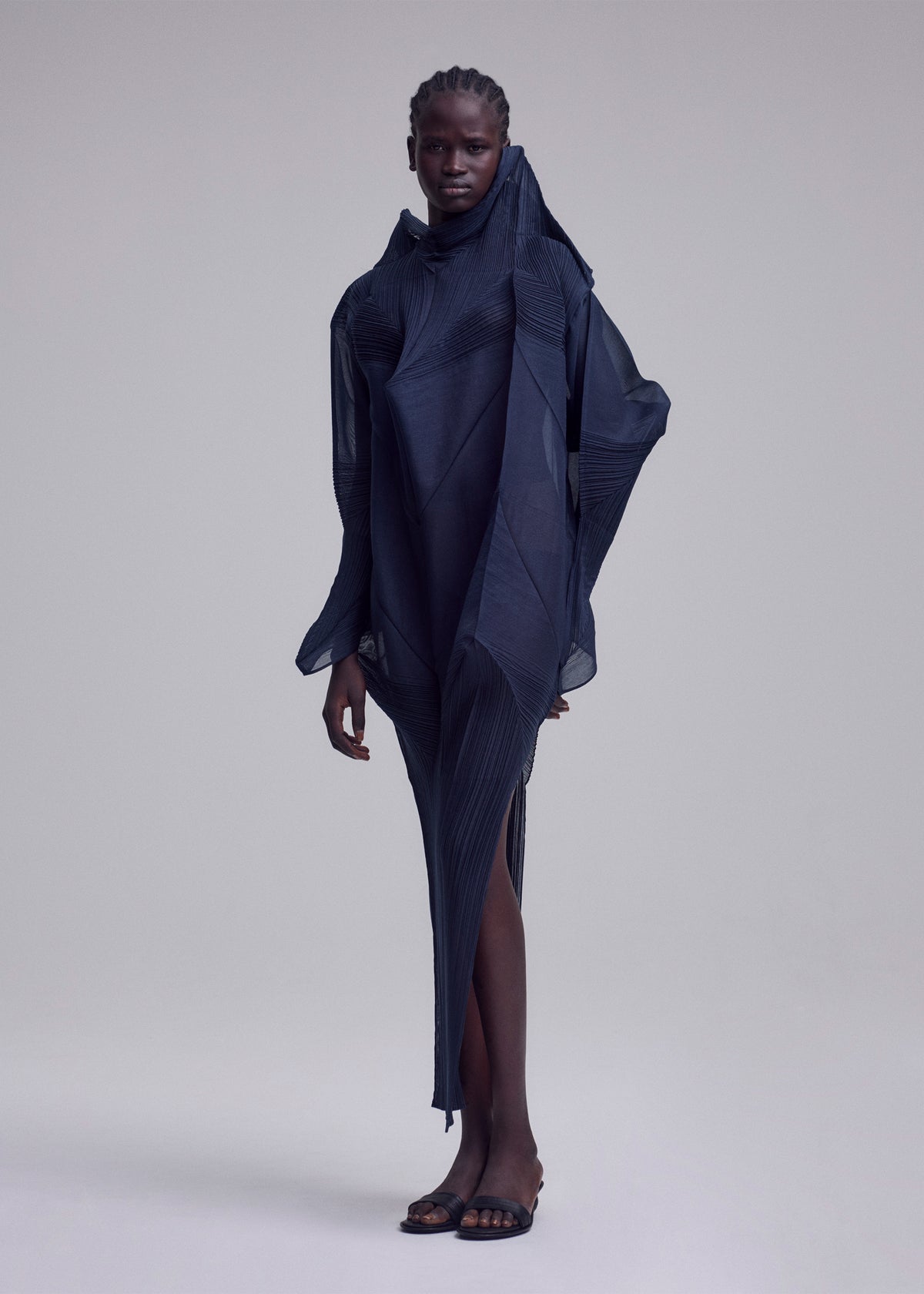 A person stands against a plain background wearing the ISSEY MIYAKE SHADE AND SHADED_NY dress, a sculptural, asymmetrical, dark navy piece with textured layers. They have short cornrowed hair and black open-toe sandals. view 10 of 10