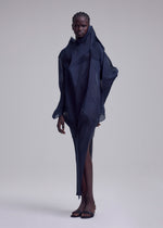 A person stands against a plain background wearing the ISSEY MIYAKE SHADE AND SHADED_NY dress, a sculptural, asymmetrical, dark navy piece with textured layers. They have short cornrowed hair and black open-toe sandals.