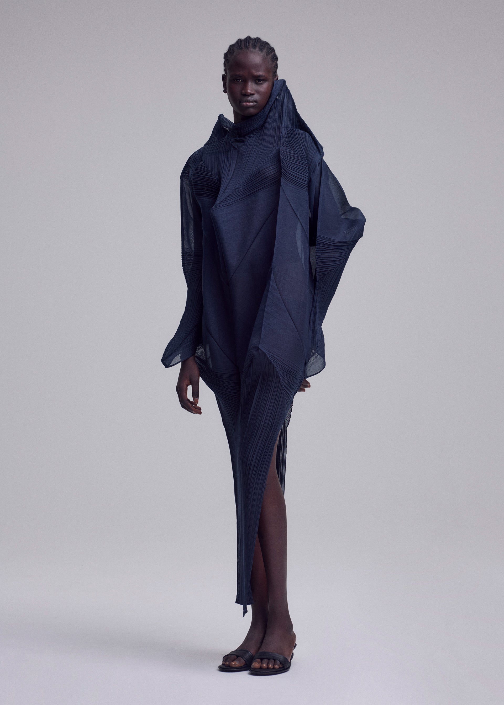 Zoomed view A person stands against a plain background wearing the ISSEY MIYAKE SHADE AND SHADED_NY dress, a sculptural, asymmetrical, dark navy piece with textured layers. They have short cornrowed hair and black open-toe sandals.