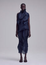 A person stands facing forward in the ISSEY MIYAKE SHADE AND SHADED_NY, a sleeveless dark blue sculptural dress with layered sheer geometric fabric, paired with open-toed black shoes, against a plain light gray background.