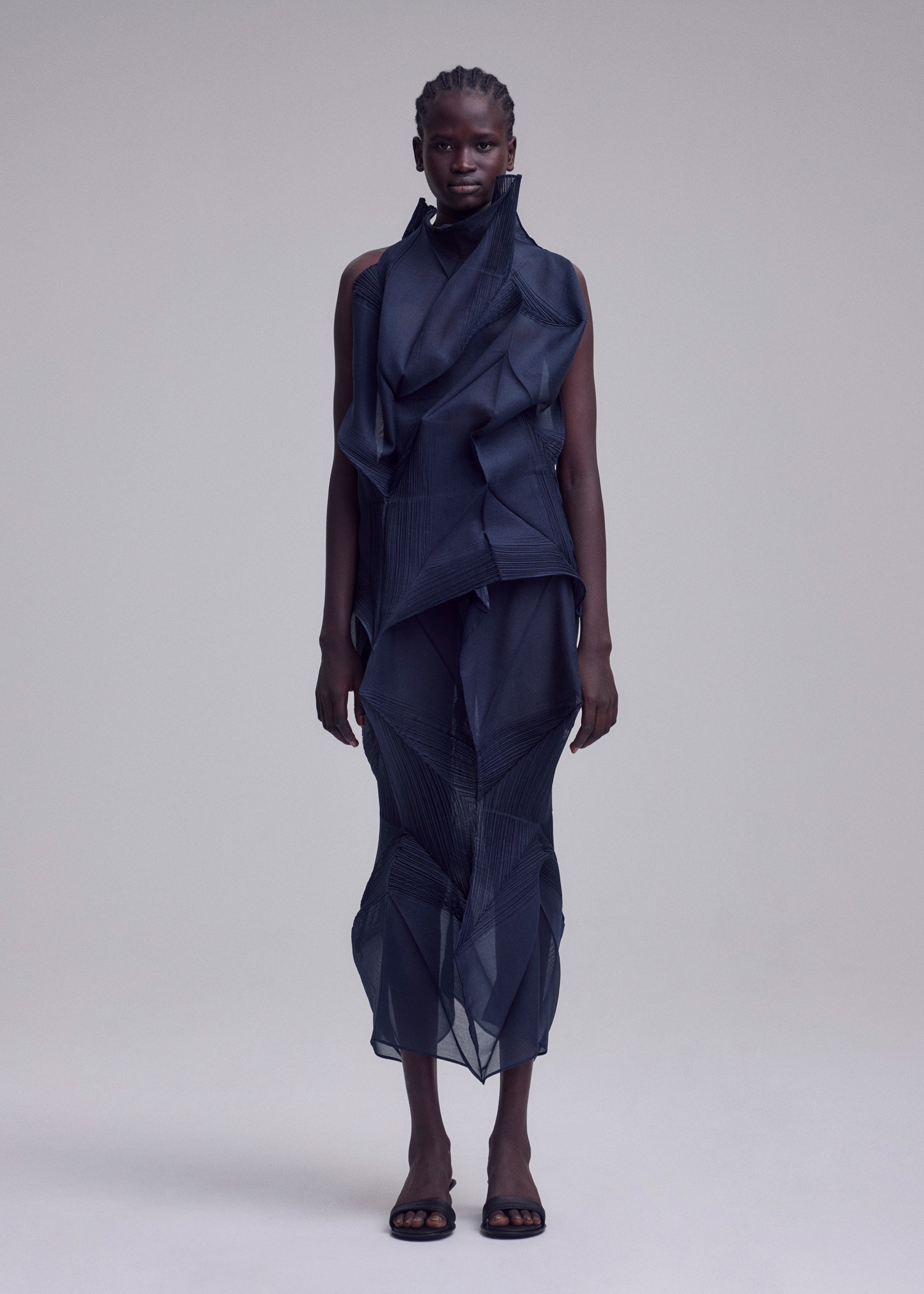 Zoomed view A person stands facing forward in the ISSEY MIYAKE SHADE AND SHADED_NY, a sleeveless dark blue sculptural dress with layered sheer geometric fabric, paired with open-toed black shoes, against a plain light gray background.