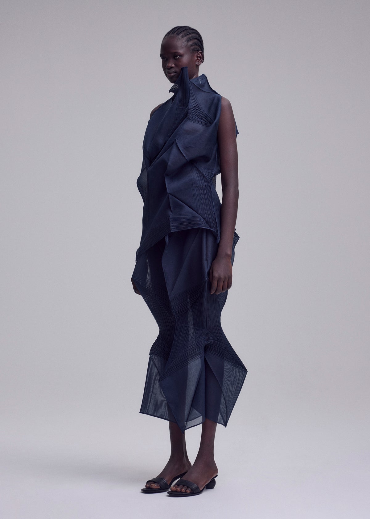 A person stands in a studio wearing the SHADE AND SHADED_NY dress by ISSEY MIYAKE, featuring geometric machine-pleated sheer dark blue layers and angular shapes, paired with open-toe black heels against a plain light background. view 4 of 11