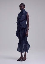 A person stands in a studio wearing the SHADE AND SHADED_NY dress by ISSEY MIYAKE, featuring geometric machine-pleated sheer dark blue layers and angular shapes, paired with open-toe black heels against a plain light background.