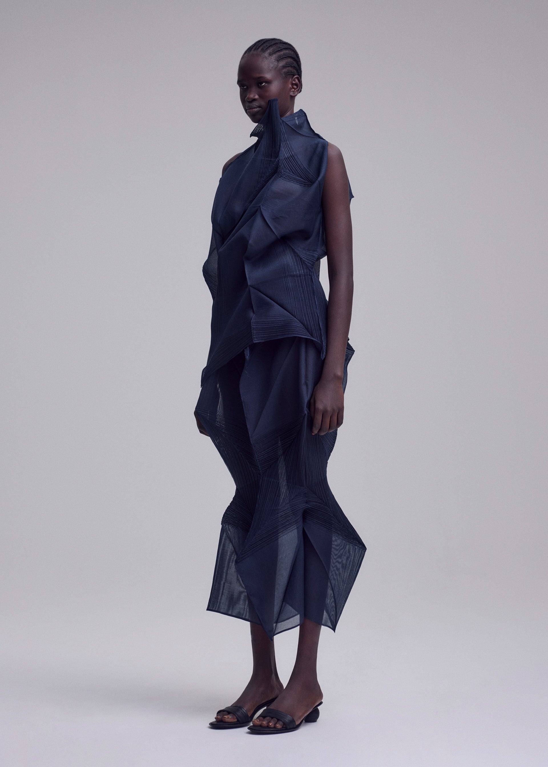 Zoomed view A person stands in a studio wearing the SHADE AND SHADED_NY dress by ISSEY MIYAKE, featuring geometric machine-pleated sheer dark blue layers and angular shapes, paired with open-toe black heels against a plain light background.