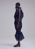 A person stands in profile against a plain background wearing ISSEY MIYAKE’s SHADE AND SHADED_NY, a dark, sheer, mid-calf sculptural silhouette with layered textures and machine-pleated details, paired with open-toe heeled sandals.