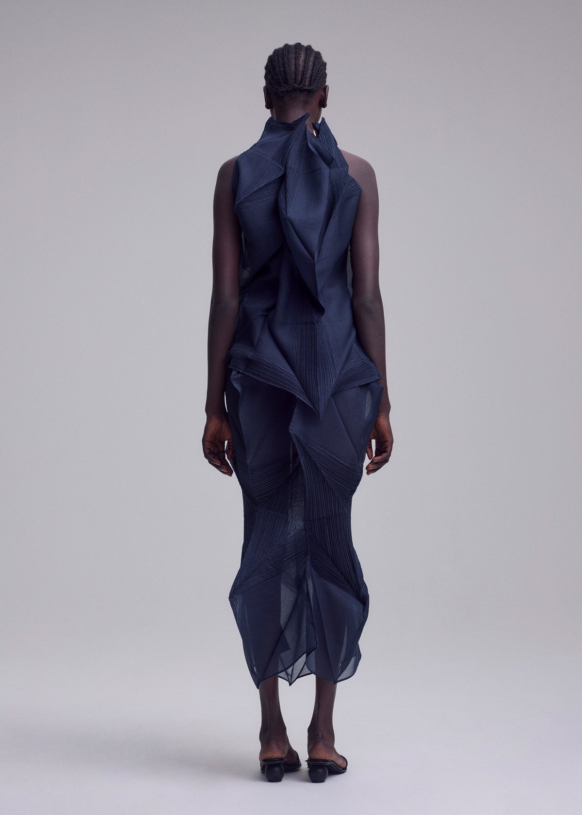 A person faces away, wearing ISSEY MIYAKE's SHADE AND SHADED_NY—a sleeveless, sculptural navy dress with geometric machine-pleating. The sheer mid-calf dress creates a striking silhouette against a plain light gray background. view 6 of 11