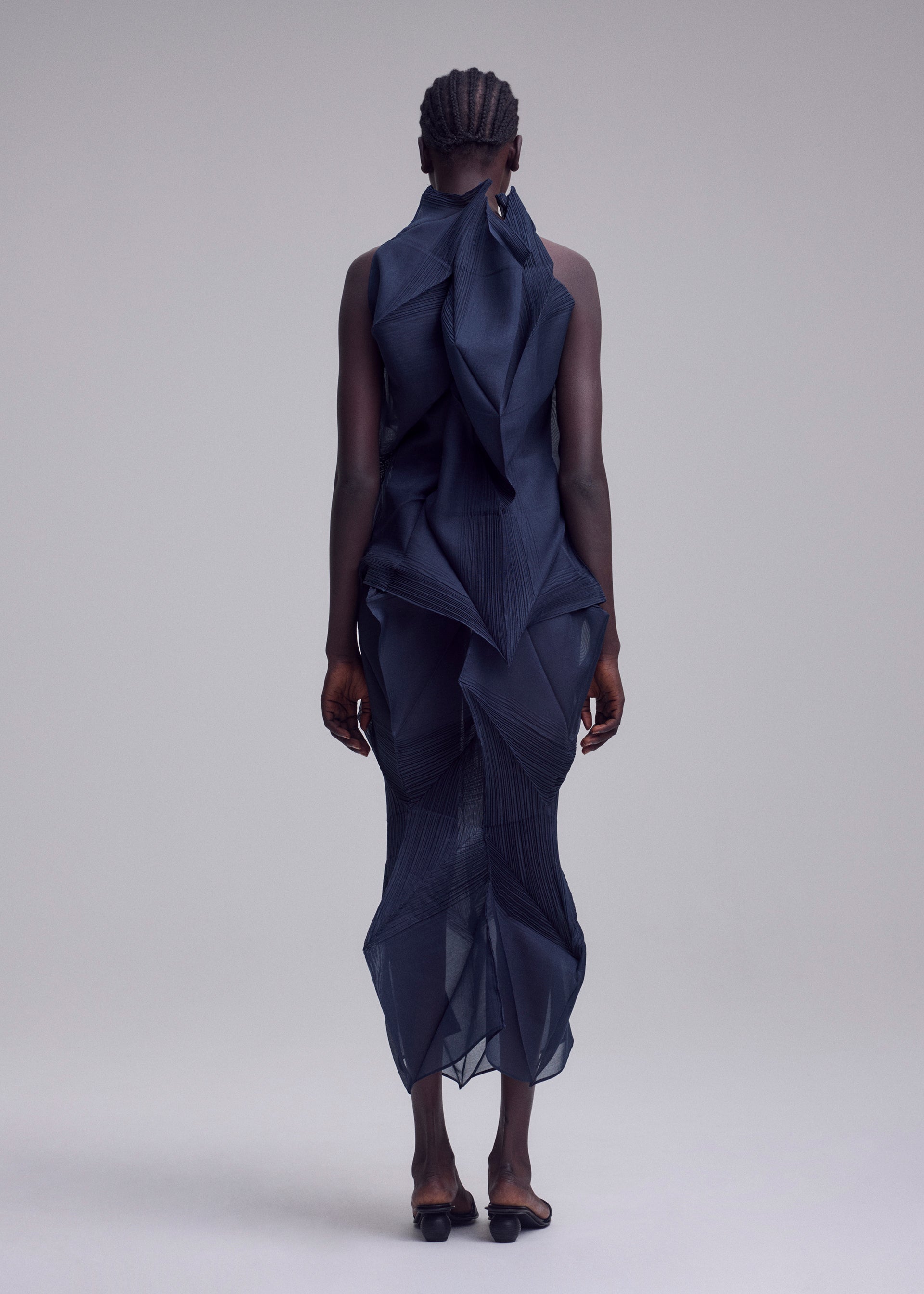 Zoomed view A person faces away, wearing ISSEY MIYAKE's SHADE AND SHADED_NY—a sleeveless, sculptural navy dress with geometric machine-pleating. The sheer mid-calf dress creates a striking silhouette against a plain light gray background.