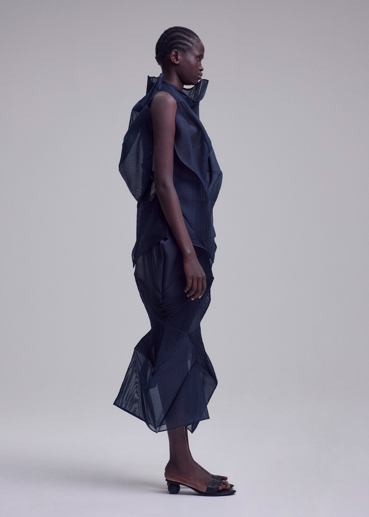 A person stands in profile against a plain background, wearing ISSEY MIYAKE’s SHADE AND SHADED_NY dress with sculptural mesh-like texture and ruffled details, paired with open-toed heeled sandals. view 7 of 11