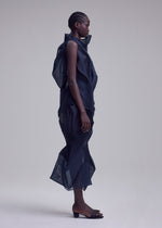 A person stands in profile against a plain background, wearing ISSEY MIYAKE’s SHADE AND SHADED_NY dress with sculptural mesh-like texture and ruffled details, paired with open-toed heeled sandals.