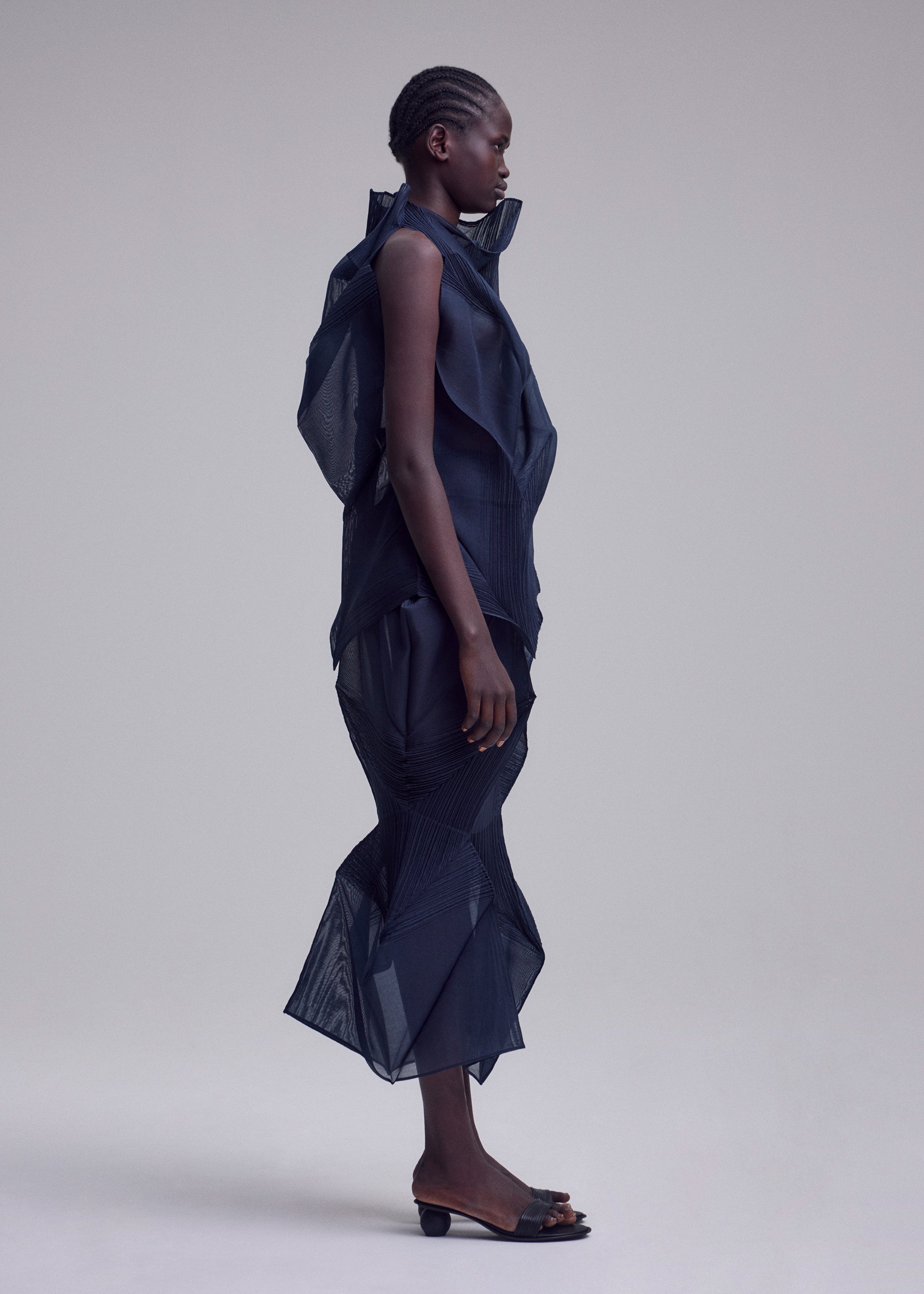 Zoomed view A person stands in profile against a plain background, wearing ISSEY MIYAKE’s SHADE AND SHADED_NY dress with sculptural mesh-like texture and ruffled details, paired with open-toed heeled sandals.