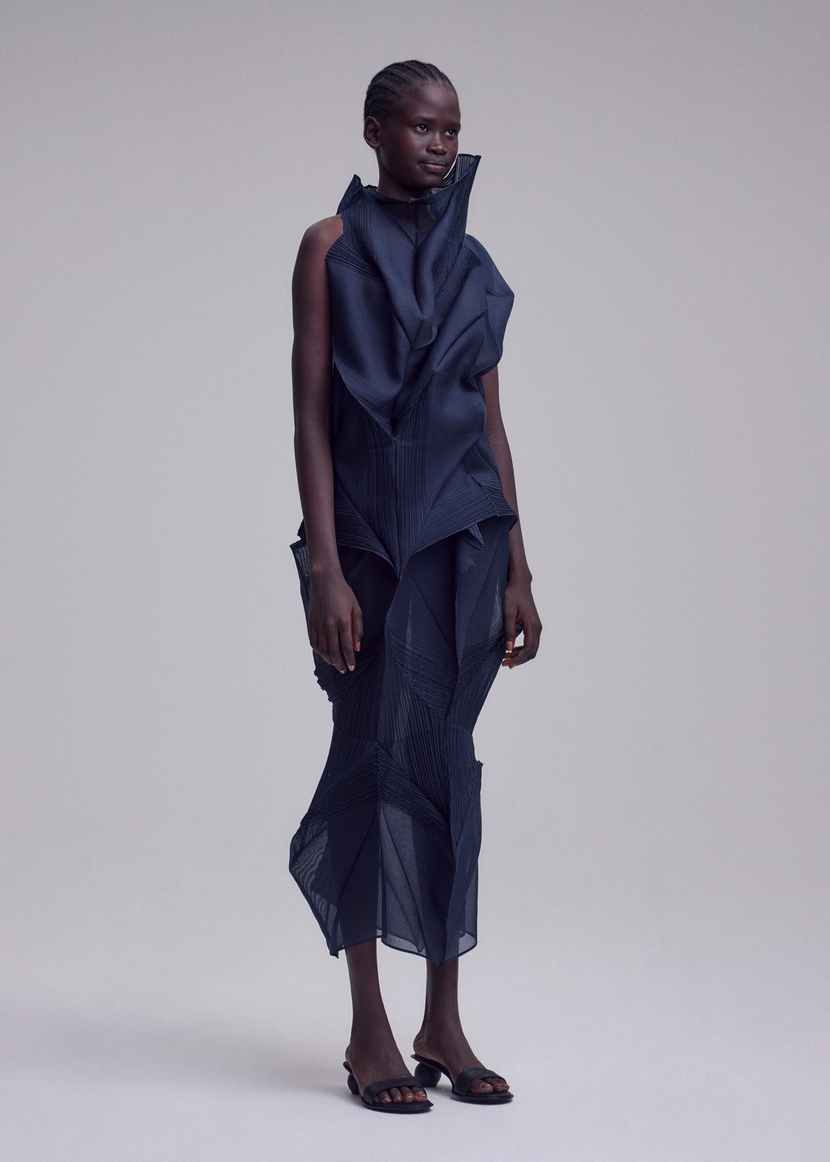 A person stands against a neutral background, wearing the SHADE AND SHADED_NY dark, machine-pleated sleeveless dress with geometric folds by ISSEY MIYAKE, styled with open-toed black shoes. view 8 of 11