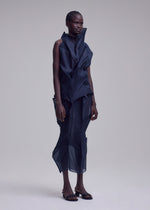 A person stands against a neutral background, wearing the SHADE AND SHADED_NY dark, machine-pleated sleeveless dress with geometric folds by ISSEY MIYAKE, styled with open-toed black shoes.