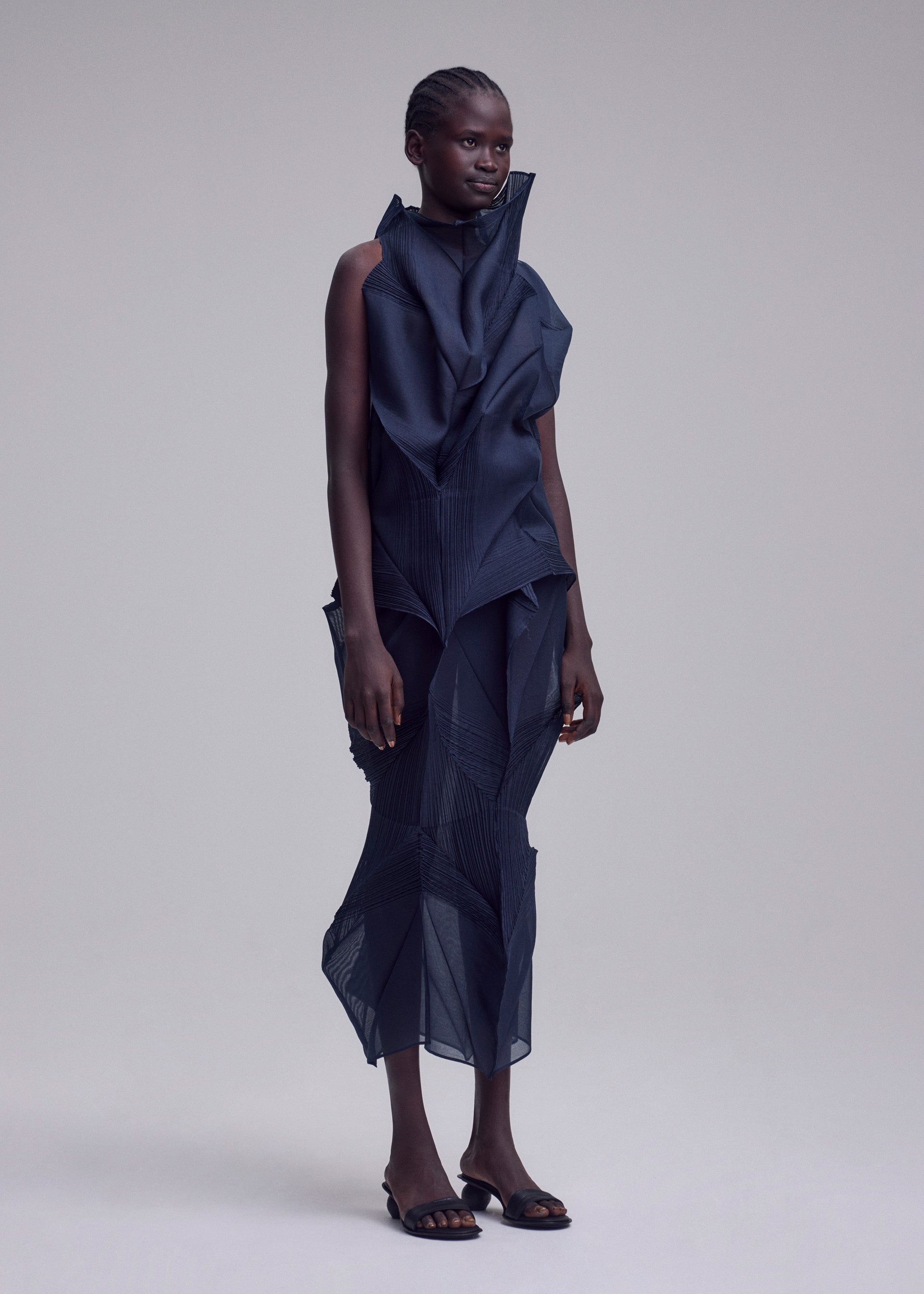 Zoomed view A person stands against a neutral background, wearing the SHADE AND SHADED_NY dark, machine-pleated sleeveless dress with geometric folds by ISSEY MIYAKE, styled with open-toed black shoes.