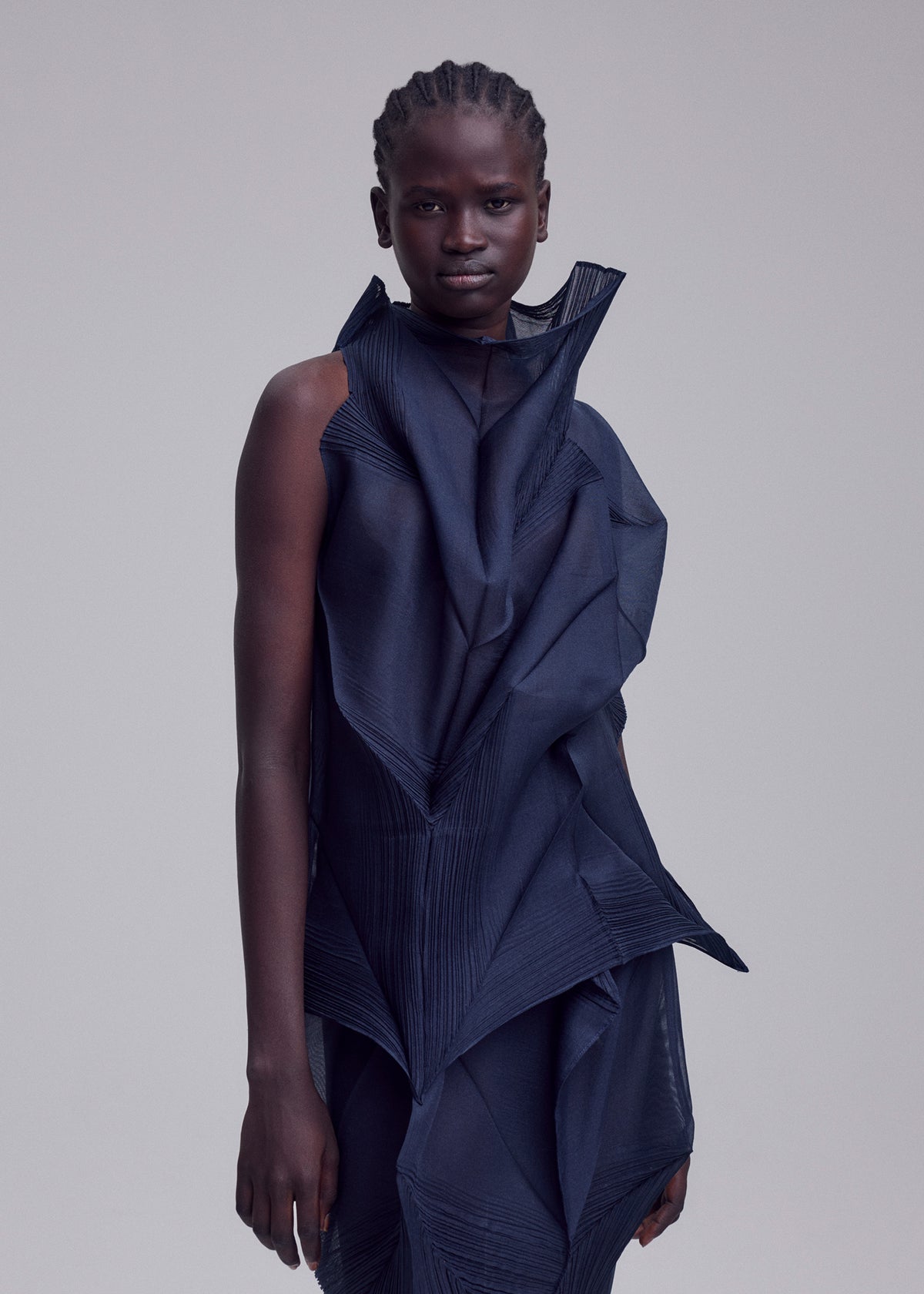 A person with braided hair models ISSEY MIYAKE's SHADE AND SHADED_NY, a dark blue sleeveless dress with machine-pleated geometric folds and a sculptural silhouette, standing against a plain light grey background. view 9 of 11