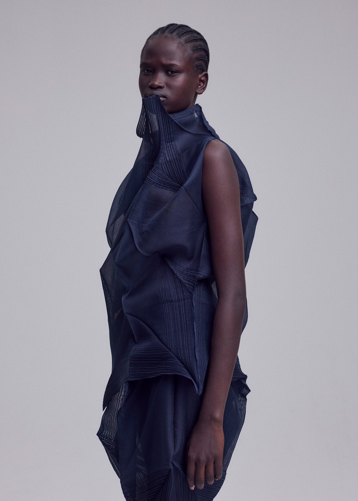 A person with dark skin and tightly braided hair wears the SHADE AND SHADED_NY sculptural, machine-pleated, semi-sheer dress by ISSEY MIYAKE. They stand against a plain light gray background, looking calmly at the camera. view 10 of 11
