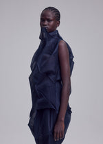 A person with dark skin and tightly braided hair wears the SHADE AND SHADED_NY sculptural, machine-pleated, semi-sheer dress by ISSEY MIYAKE. They stand against a plain light gray background, looking calmly at the camera.