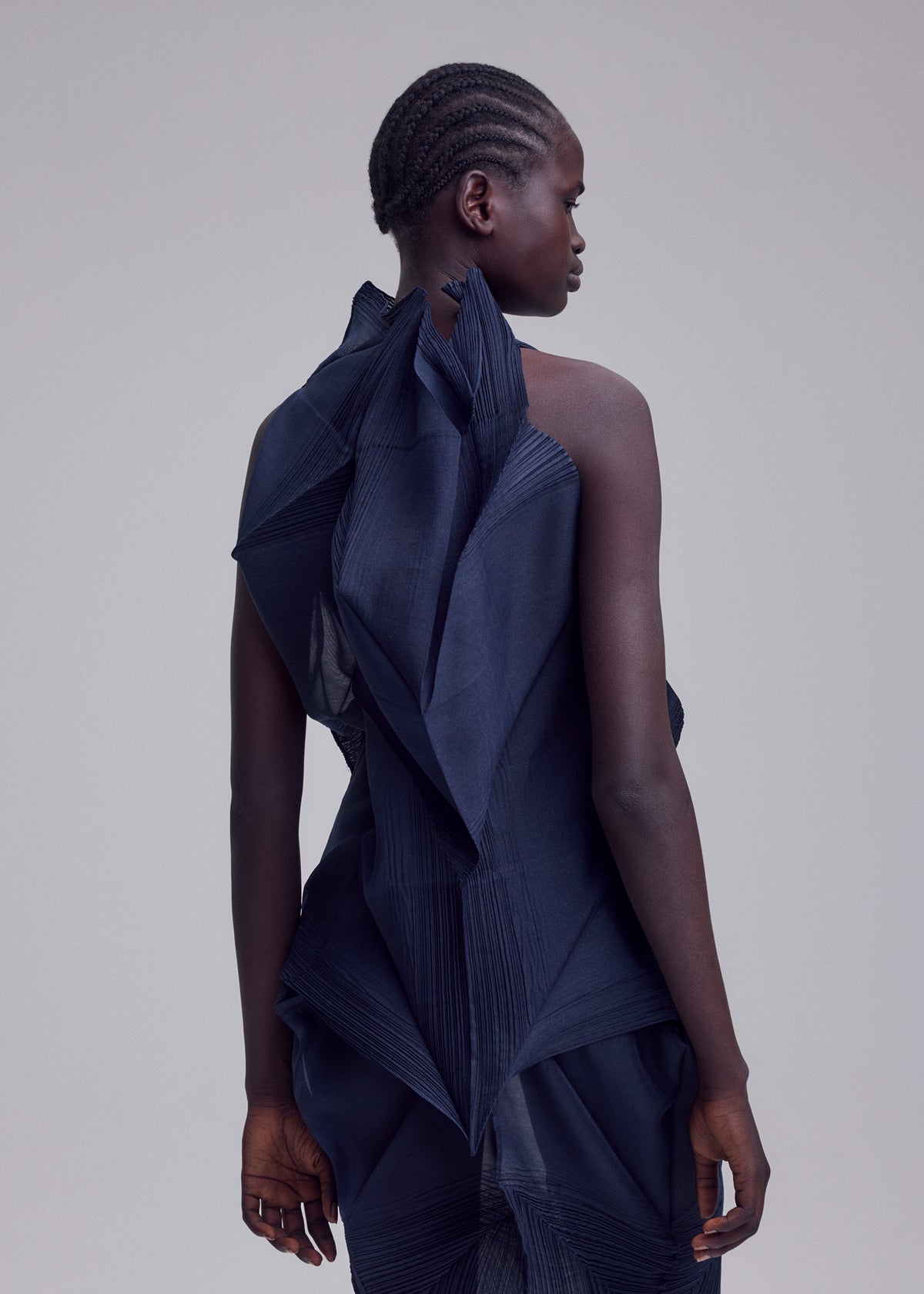 A person with short, braided hair faces away, wearing the SHADE AND SHADED_NY dress by ISSEY MIYAKE—a dark, sculptural piece with machine-pleated, angular details—set against a light background. view 11 of 11