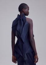 A person with short, braided hair faces away, wearing the SHADE AND SHADED_NY dress by ISSEY MIYAKE—a dark, sculptural piece with machine-pleated, angular details—set against a light background.