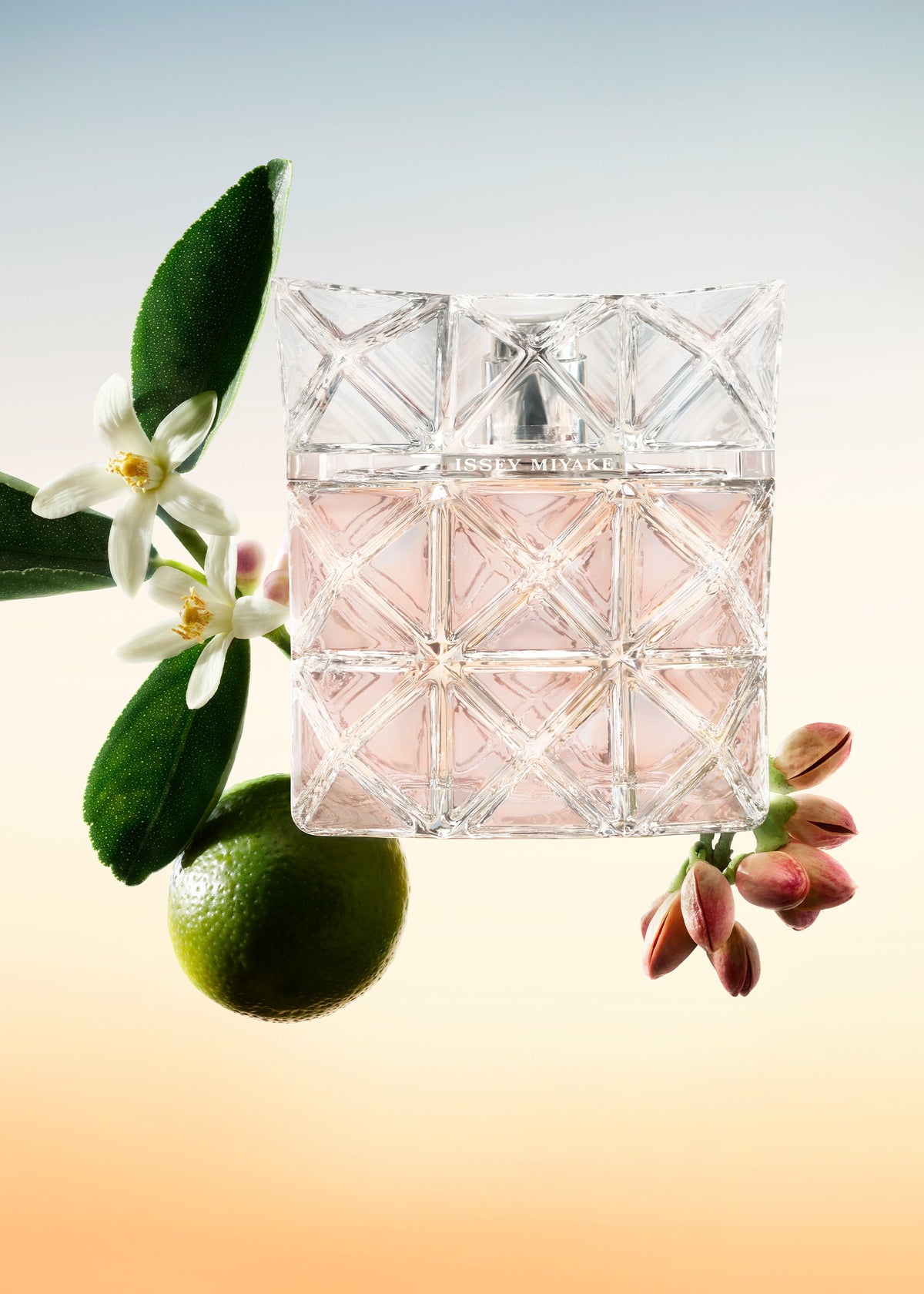A crystal-patterned bottle of LUMIERE D’ISSEY EAU DE PARFUM by ISSEY MIYAKE PARFUMS, filled with pink liquid, is surrounded by white flowers, green leaves, a whole lime, and reddish buds against a soft gradient background. view 3 of 3