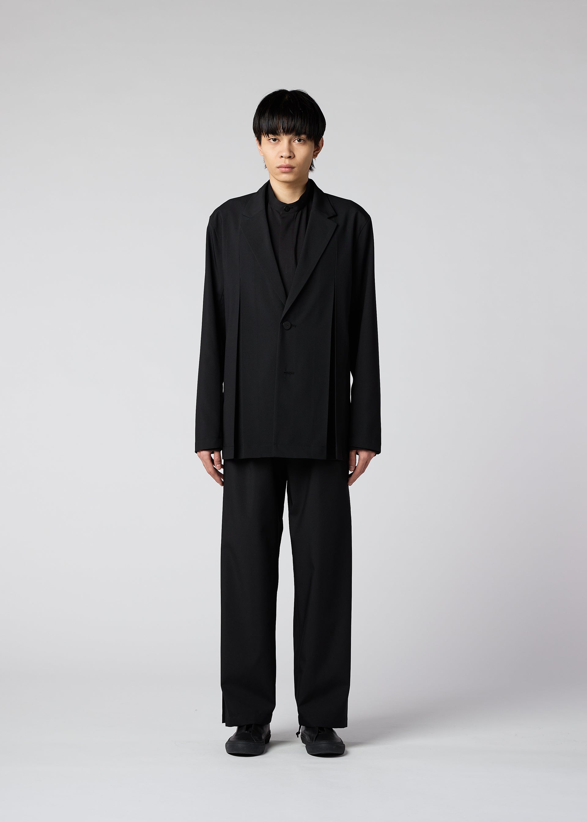 Zoomed view A person with short black hair faces forward, wearing the WINGS oversized black suit with wide handmade-pleated pants from IM MEN and black shoes, set against a plain light gray background.