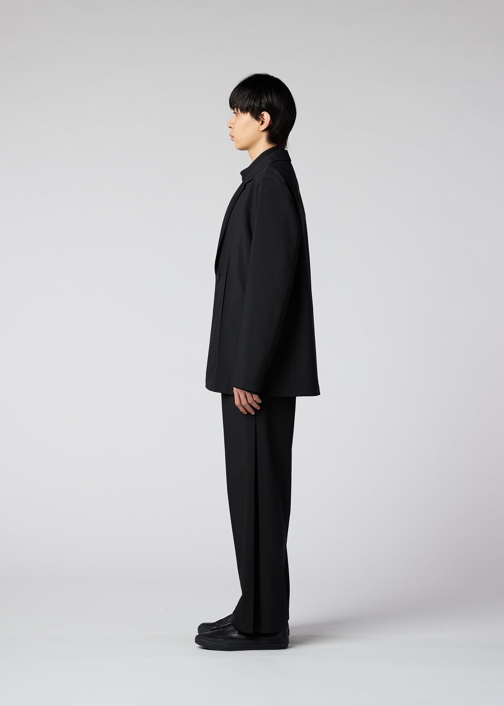 Zoomed view A person with straight black hair stands in profile against a plain light gray background, wearing the IM MEN WINGS loose-fitting black suit with wide pants and black shoes.