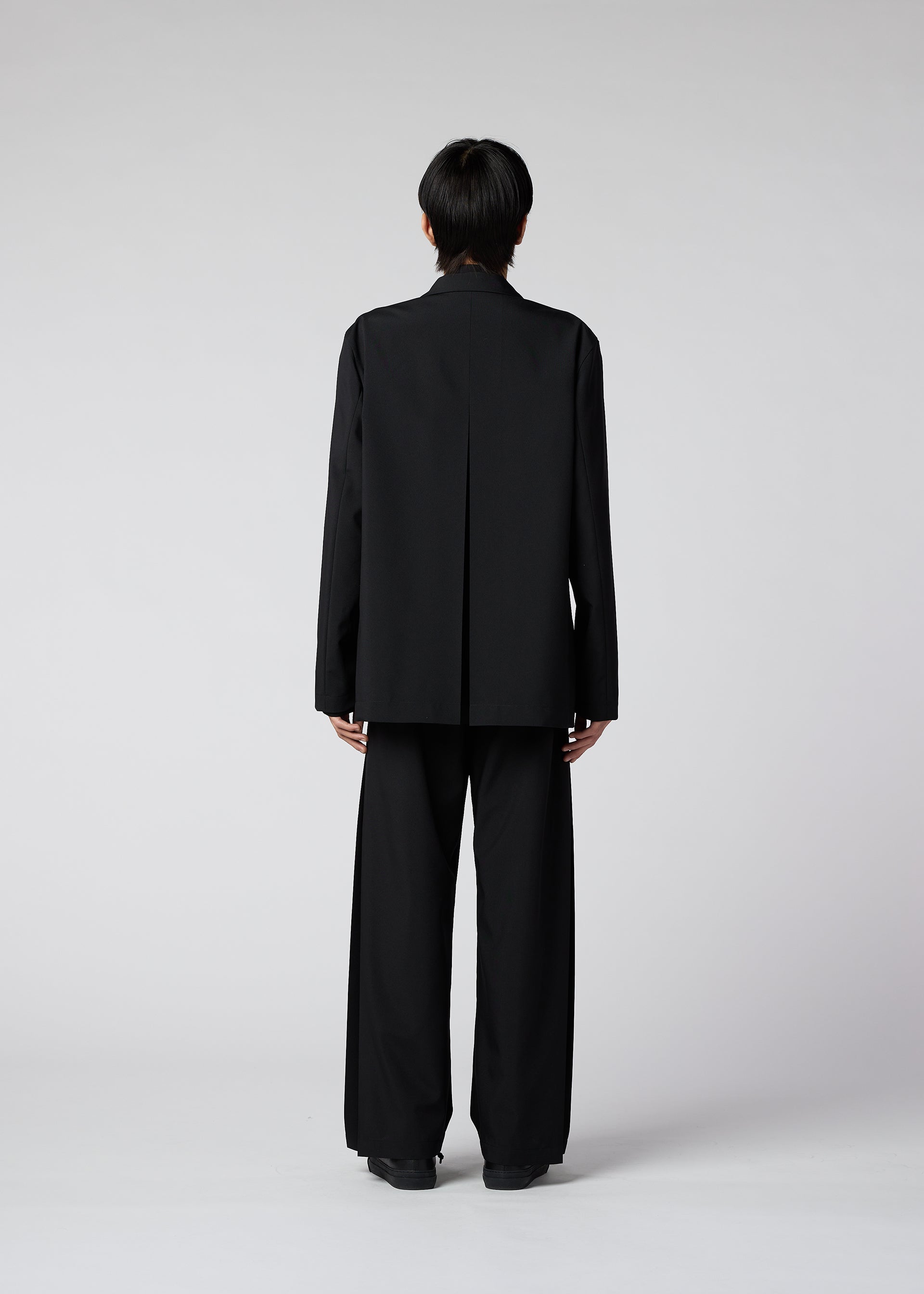 Zoomed view Someone with short dark hair faces away from the camera, wearing the IM MEN WINGS loose black suit with wide pants and black shoes, set against a plain light gray background.