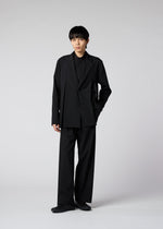 A young person models the IM MEN WINGS suit—loose-fitting black with wide pants and handmade pleats—paired with a black shirt and shoes, hand in pocket, embodying effortless style and easy care against a light gray backdrop.