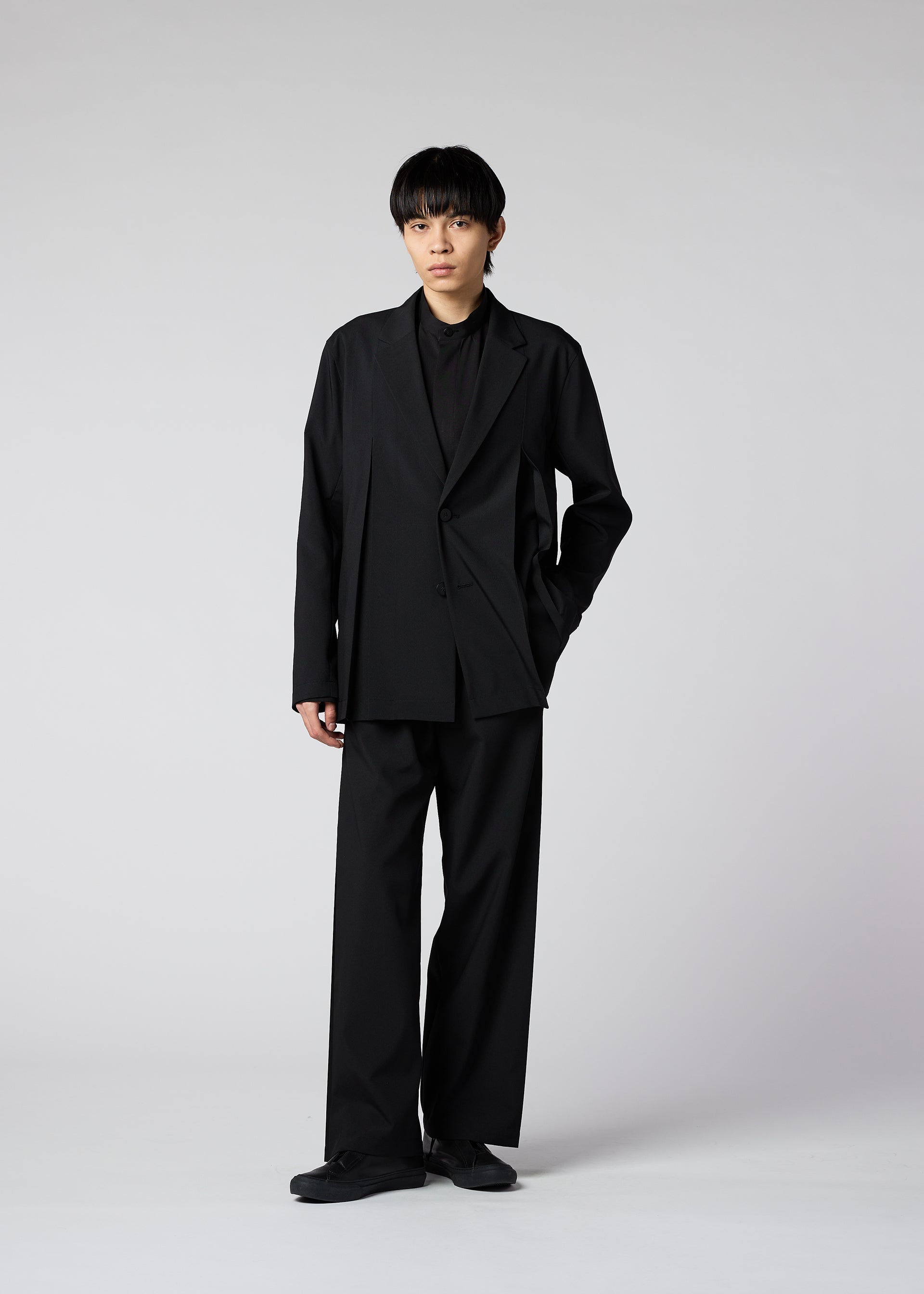 Zoomed view A young person models the IM MEN WINGS suit—loose-fitting black with wide pants and handmade pleats—paired with a black shirt and shoes, hand in pocket, embodying effortless style and easy care against a light gray backdrop.