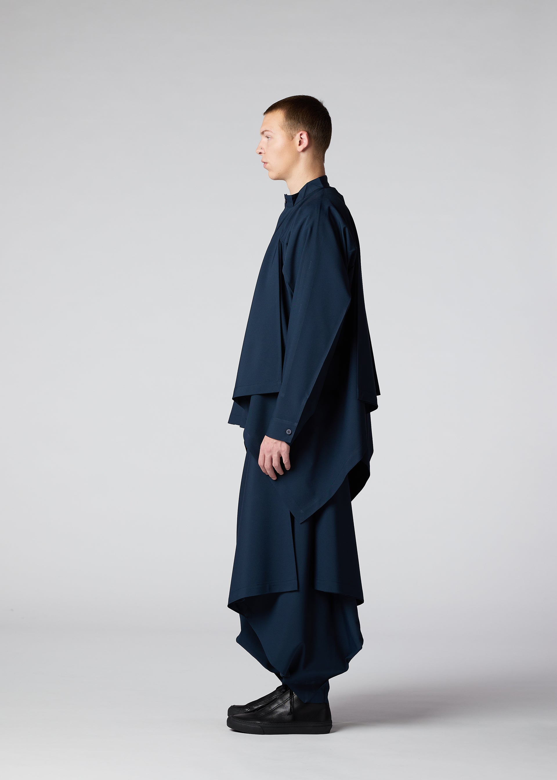 Zoomed view A person stands in profile against a plain light background, wearing the IM MEN FLAT DRAPE set—loose-fitting, plant-based polyester with pleated details and voluminous navy blue draping on wide sleeves and pant legs—paired with black shoes.