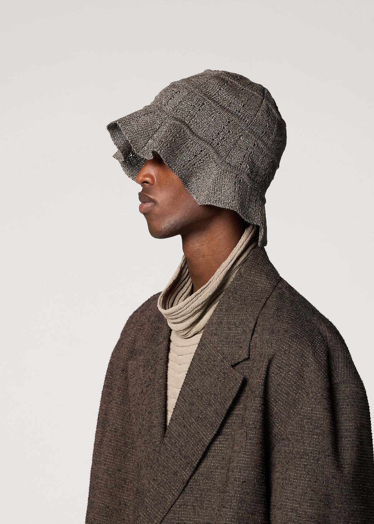 A person in a textured brown coat and beige turtleneck faces left, wearing the gray ENGRAVE HAT by IM MEN, which features wide-brimmed waved patterns inspired by ceramic artist Shoji Kamoda. The background is plain and light-colored. view 3 of 3